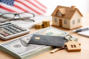 Realistic and minimalist composition showing consumer finances in the United States, with a credit card resting on a stack of US dollar bills, house keys attached to a wooden home-shaped keychain, and a small model house in the background, suggesting financial decisions influenced by interest rates and access to credit.